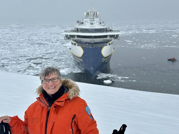 Sheila Gallant-Halloran stands on a snowy Antarctic shoreline in expedition gear, smiling with a trekking pole in hand after hiking up an icy hill to see penguins. In the background, a zodiac lands guests near the shore while the expedition ship National Geographic Endurance sits anchored among dramatic Antarctic scenery.