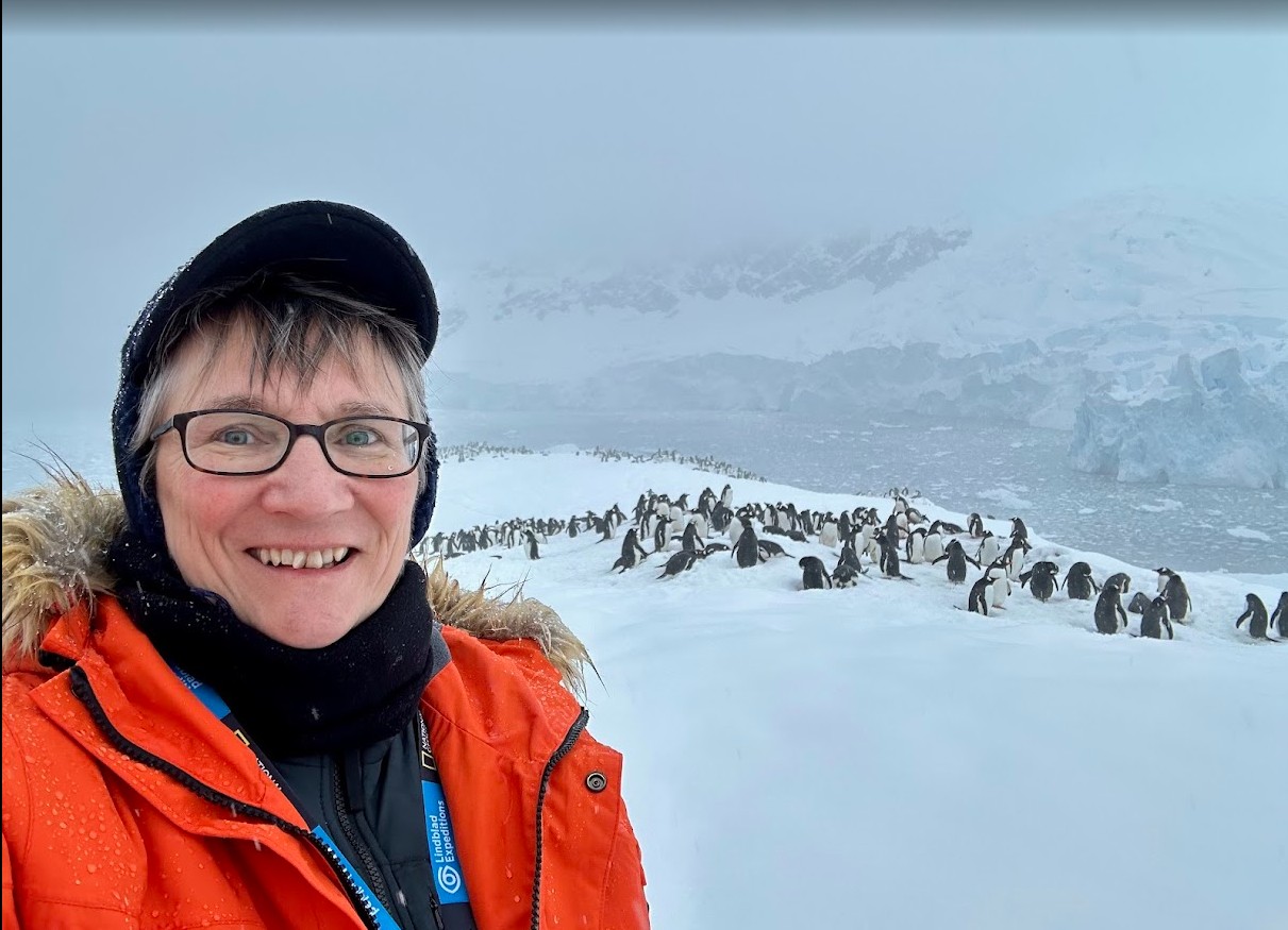 Sheila Gallant-Halloran standing at Neko Harbour, Antarctica, in front of a penguin colony with snow-covered glaciers and icy mountains in the background.